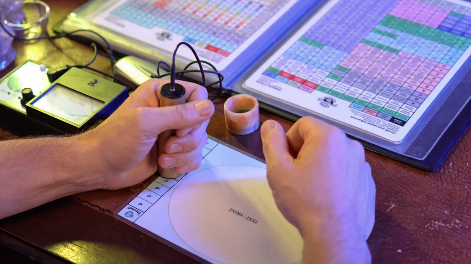Hands holding a pendulum-like stick over a printed dial chart beside reference rate tables on a wooden desk